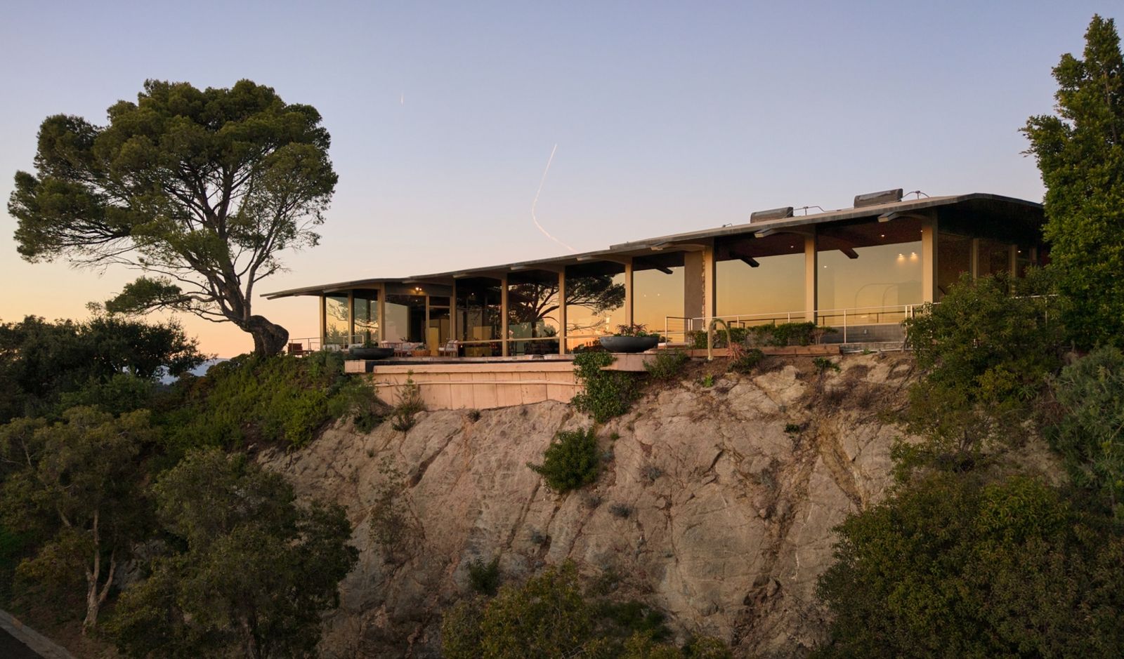 Moderne, eingeschossige Villa auf einem felsigen Hang. Große Glasfronten öffnen den Blick ins Innere des Hauses, umgeben von Bäumen und Sträuchern im Licht der Dämmerung. 29-624_Cole_Pl_-_Nils_Timm.jpg