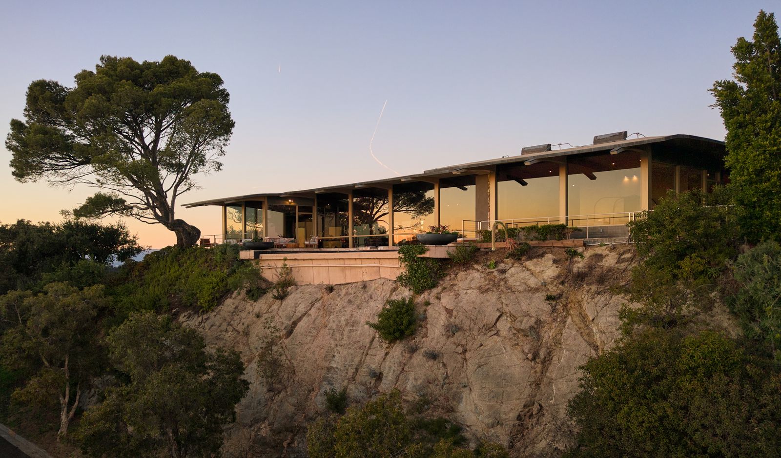 Moderne, eingeschossige Villa auf einem felsigen Hang. Große Glasfronten öffnen den Blick ins Innere des Hauses, umgeben von Bäumen und Sträuchern im Licht der Dämmerung. 29-624_Cole_Pl_-_Nils_Timm.jpg