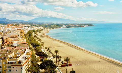 Weitläufiger Panoramablick auf einen langen Sandstrand an der Costa del Sol. Entlang der Küste reihen sich Wohnhäuser, Hotels und Palmen aneinander. Das türkisfarbene Mittelmeer erstreckt sich bis zum Horizont, während im Hintergrund Berge unter einem leicht bewölkten Himmel aufragen. 1500x900_AdobeStock_1488407459.jpg