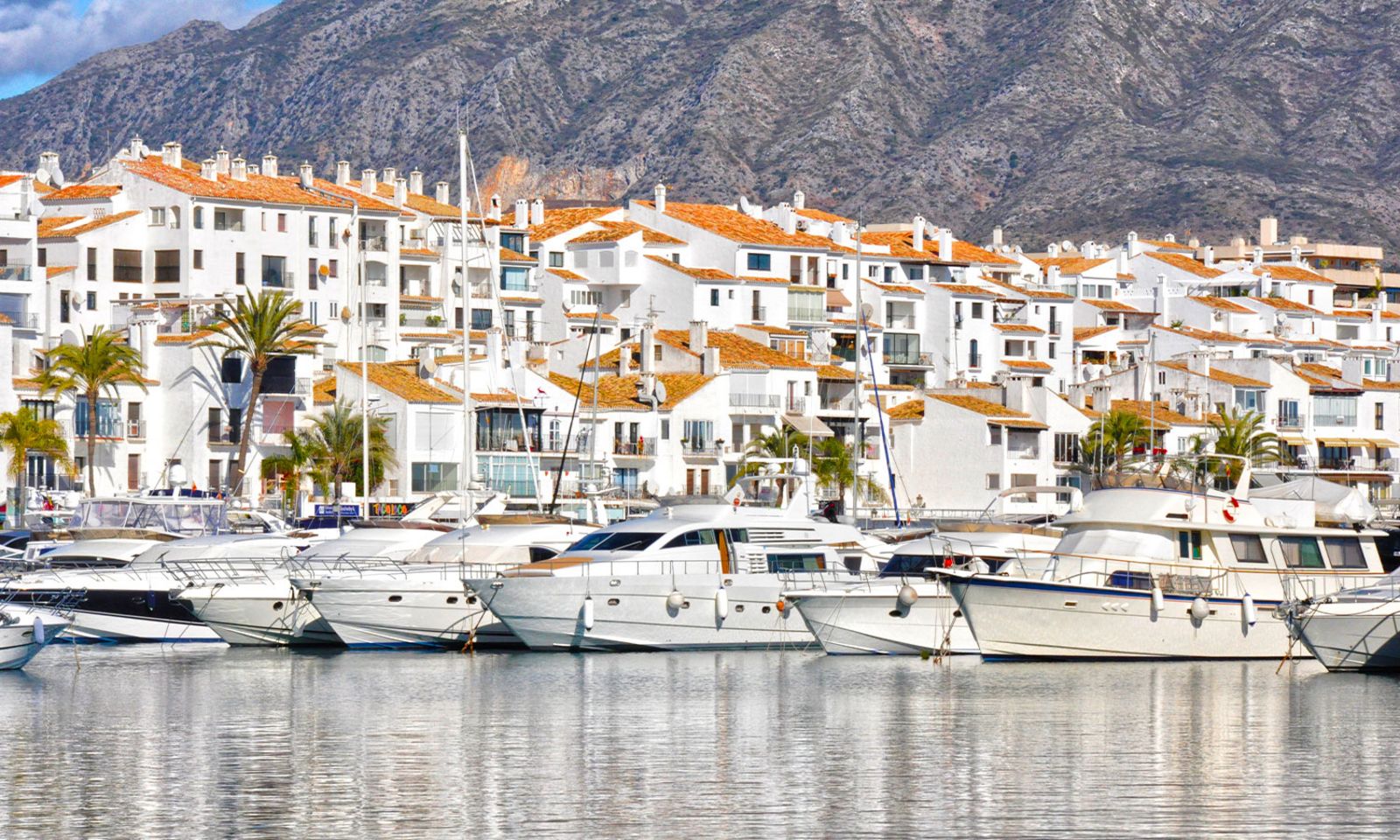 Mehrere luxuriöse weiße Yachten liegen im Hafen von Puerto Banús an der Costa del Sol. Im Hintergrund stehen dicht aneinandergereihte weiße Gebäude mit terrakottafarbenen Dächern, Palmen und einer imposanten Bergkulisse. 1500x900_AdobeStock_77572572jpg.jpg