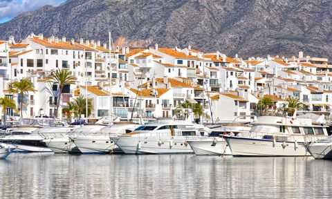 Mehrere luxuriöse weiße Yachten liegen im Hafen von Puerto Banús an der Costa del Sol. Im Hintergrund stehen dicht aneinandergereihte weiße Gebäude mit terrakottafarbenen Dächern, Palmen und einer imposanten Bergkulisse. 1500x900_AdobeStock_77572572jpg.jpg