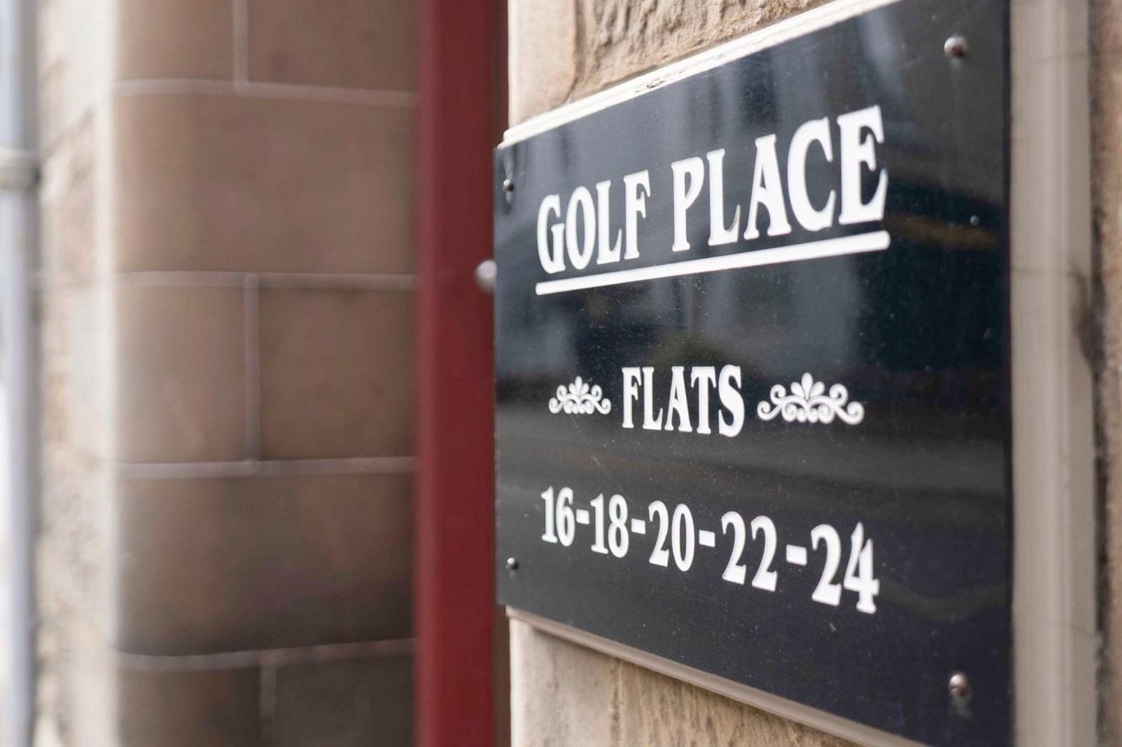 Schwarzes Gebäudeschild mit der Aufschrift „Golf Place Flats“ und den Wohnungsnummern 16 bis 24 an einer Steinfassade. 53151482.jpg