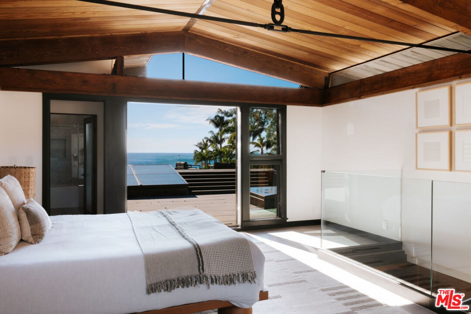 Schlafzimmer mit Bett vor einer Treppe ins Untergeschoss und weit geöffneter Fenstertür zur Holzterrasse mit Meerblick. origin7.jpg