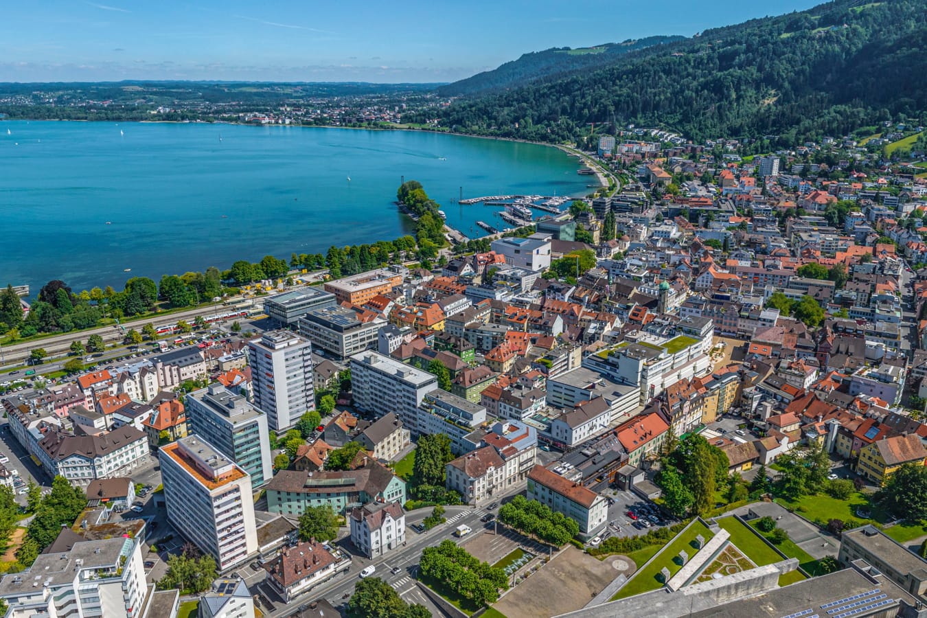 Bodensee, Luftaufnahme einer Stadt am türkisblauen See, mit dicht bebauten Ufern, Booten im Wasser und grüner Hügellandschaft im Hintergrund. AdobeStock_571802314_ARochau.jpeg