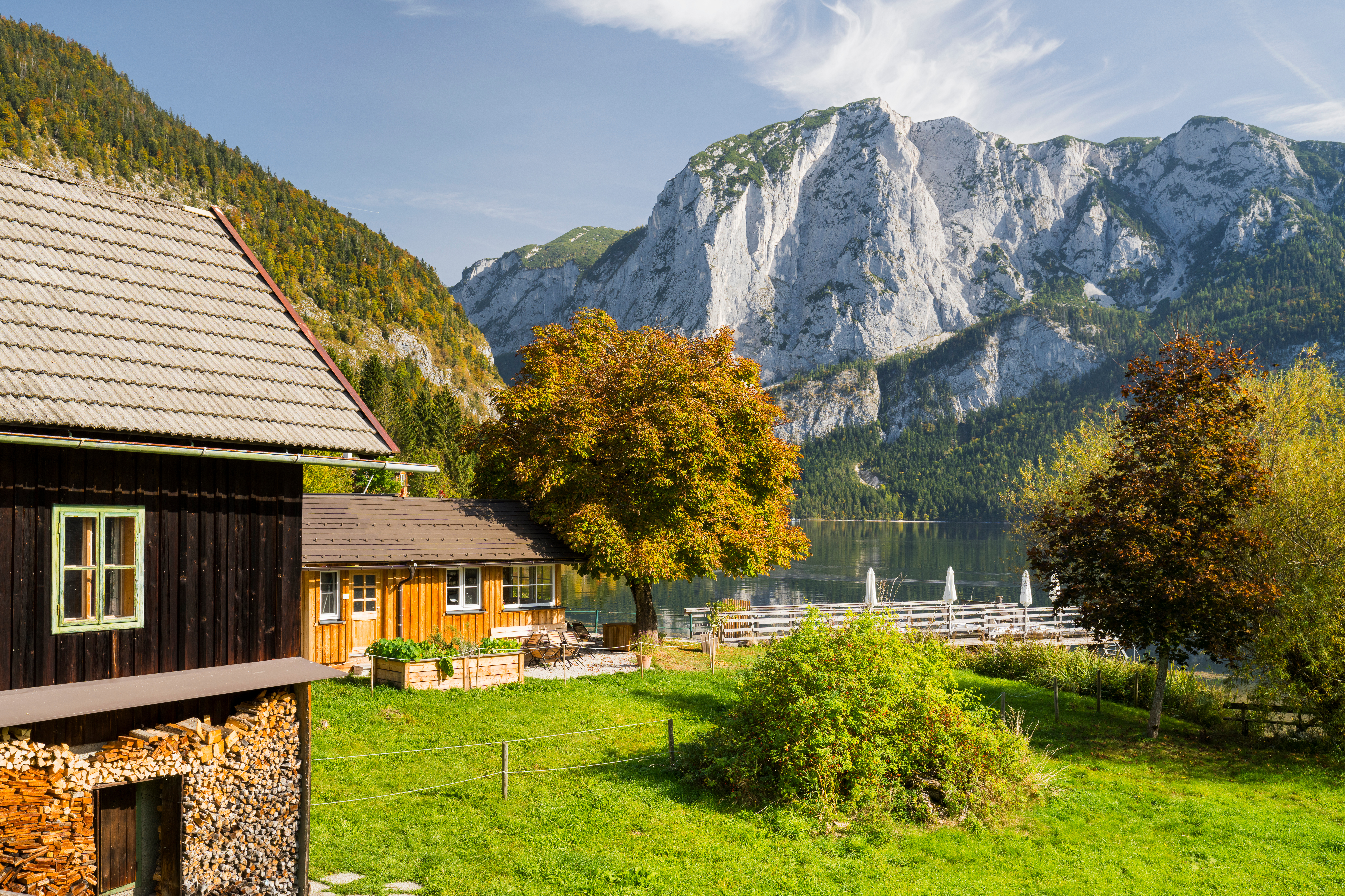 Region Salzkammergut, Ansicht Holzhaus auf einer grünen Almwiese mit Blick auf einen See und umliegende Berge unter klarem Himmel. AdobeStock_758782508_Rainer_Mirau.jpeg